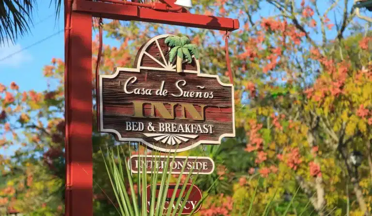 A sign for "Casa de Sueños Inn Bed & Breakfast" hanging on a red post, surrounded by flowering trees.