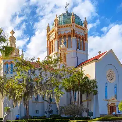 A historic, light-colored church with a green dome and cross stands against a blue sky with white clouds.