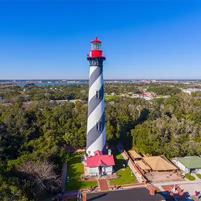A St. Augustine lighthouse with a black and white spiral pattern stands in a wooded area under a clear blue sky.