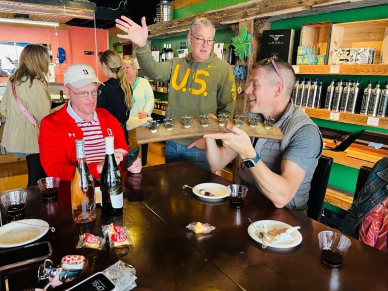 A man in a gray vest serves a flight of drinks to others seated at a wooden table in a brightly colored room.
