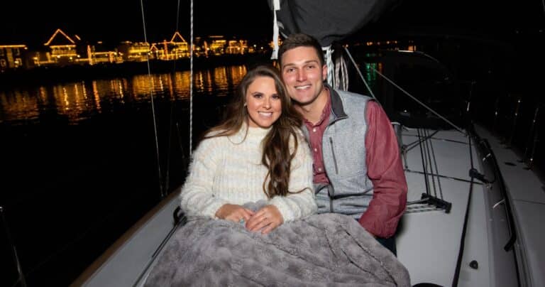 A couple sits under a blanket on a boat at night, with illuminated buildings reflecting on the water behind them.