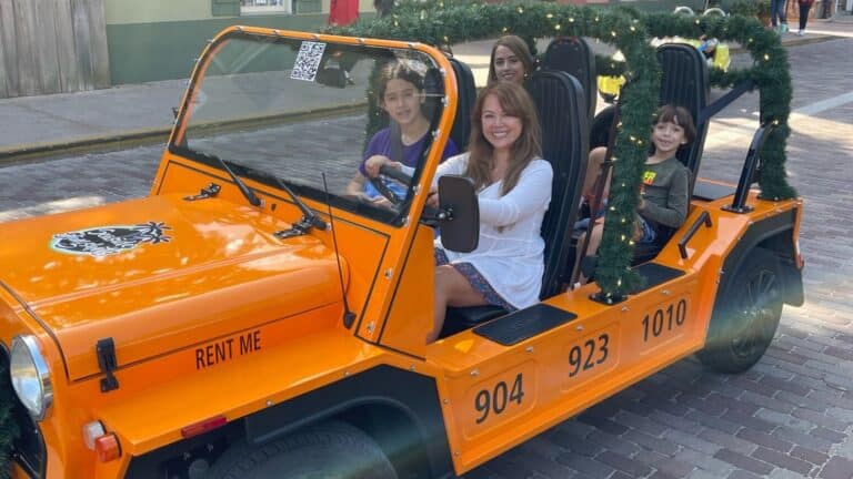 A group sits in an orange, festive open-air vehicle parked on a brick street.