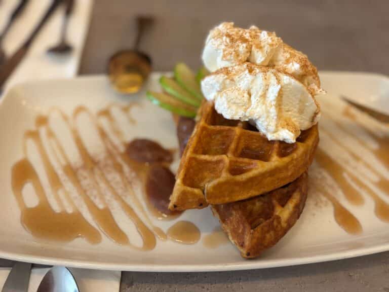 A rectangular plate holds a waffle topped with whipped cream, next to sliced green fruit and a drizzle of syrup.