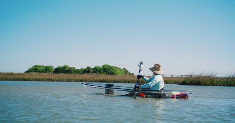 A person in a sun hat paddles a kayak through calm, marshy water under a clear blue sky.