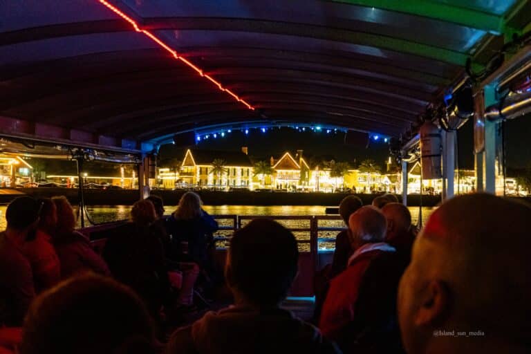 View from a boat at night, with passengers watching illuminated waterfront buildings in the distance.