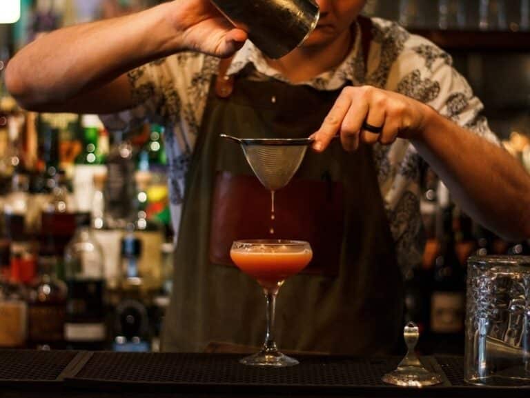 A bartender pours a cocktail through a mesh strainer into a stemmed glass at a dimly lit bar.