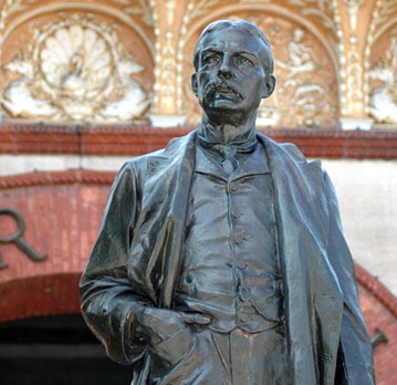 A dark bronze statue of a man in a waistcoat and coat stands in front of a building with arched, ornate masonry.