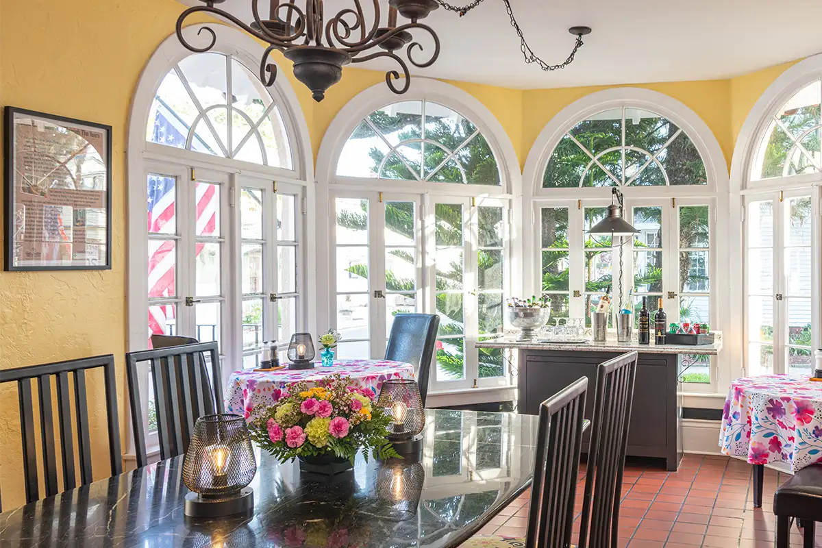 A sunny room with yellow walls, arched windows, floral-covered tables, a dining set, and a hanging chandelier.