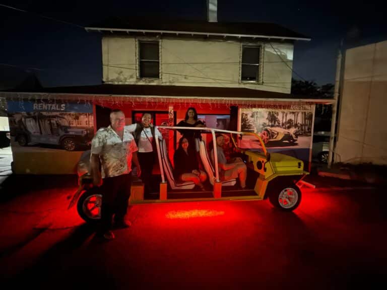 A group of people sit in a yellow open-air vehicle parked at night, illuminated by bright red ground-effect lighting.