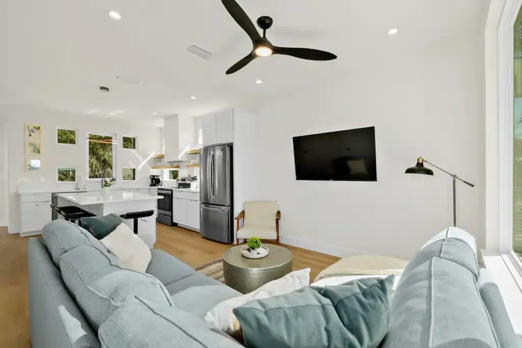 A modern living room with a light gray sofa, ceiling fan, wall-mounted TV, and open-plan kitchen in the background.