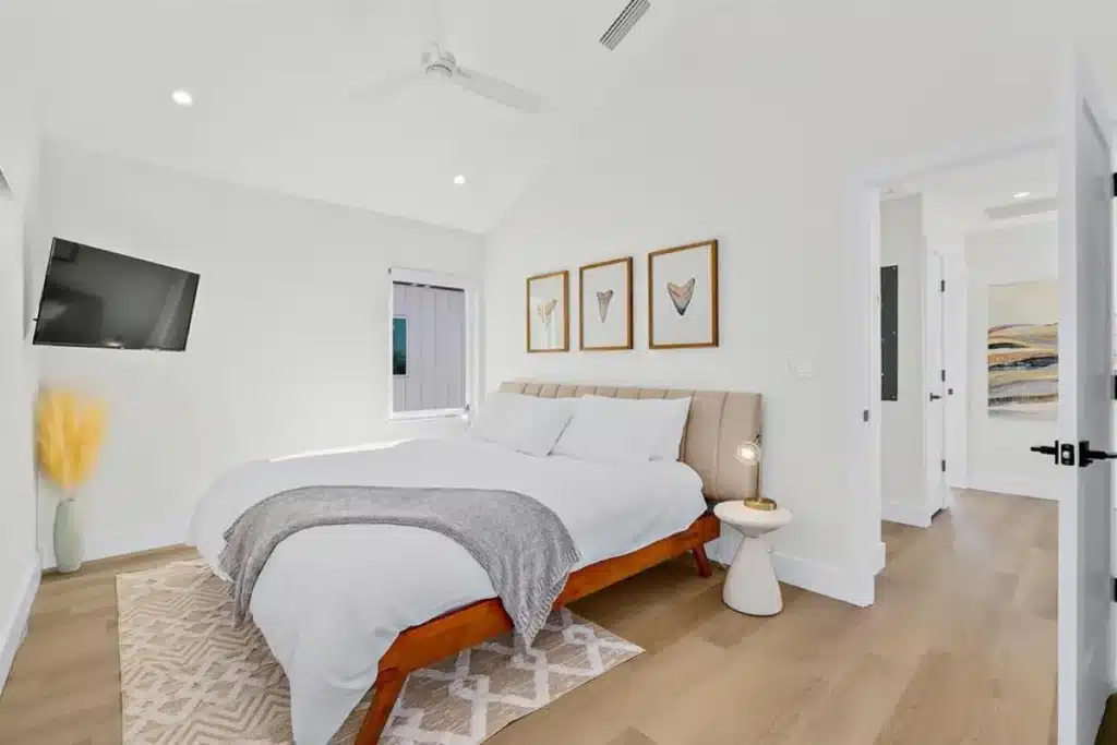 A modern bedroom with a bed, white walls, wood floors, a ceiling fan, and framed art above the headboard.