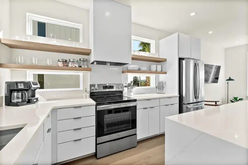 Modern kitchen with white cabinets, stainless steel appliances, open wood shelving, and a large island.