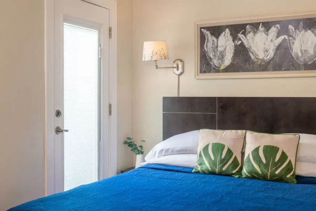 A bedroom with a blue quilt on the bed, two leaf-patterned pillows, a dark headboard, and wall art above the bed.