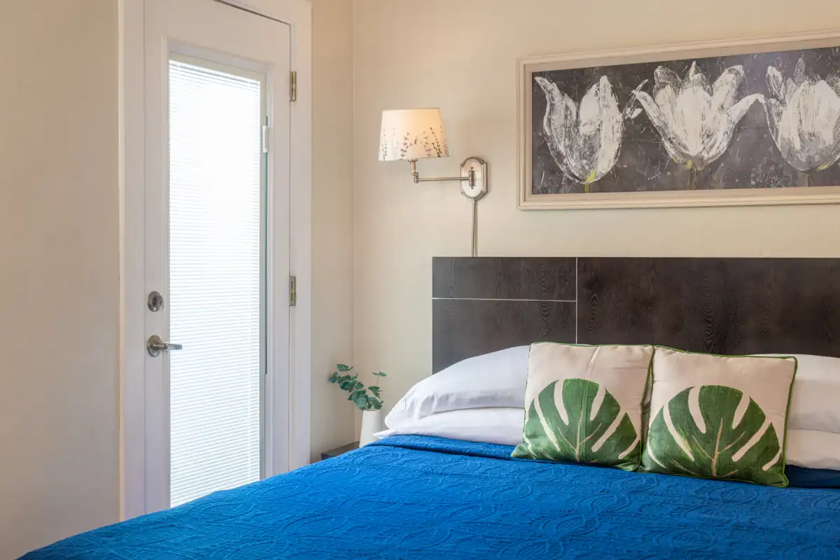 A bedroom with a blue quilt on the bed, two leaf-patterned pillows, a dark headboard, and wall art above the bed.