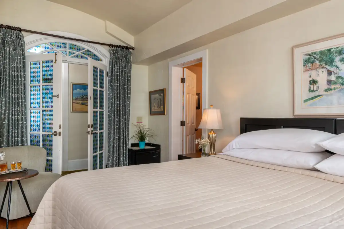 A hotel bedroom with a large bed, a small round table with glasses, glass paneled doors, and a framed wall picture.