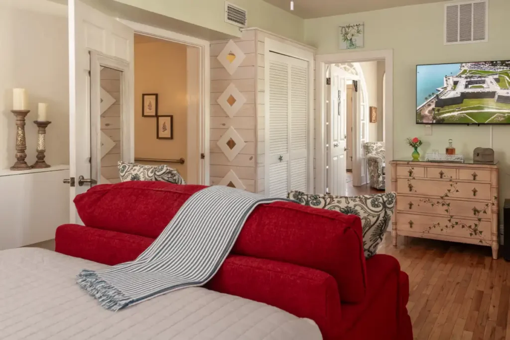 A bedroom features a bright red couch, a light-colored bed, a wooden dresser, and a mounted television.