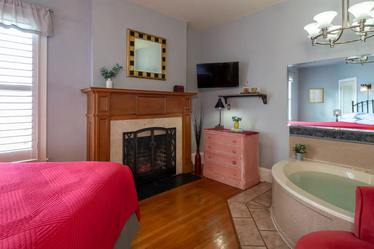 A bedroom with a red bed, a fireplace, a pink dresser, and a corner bathtub with a mirror.