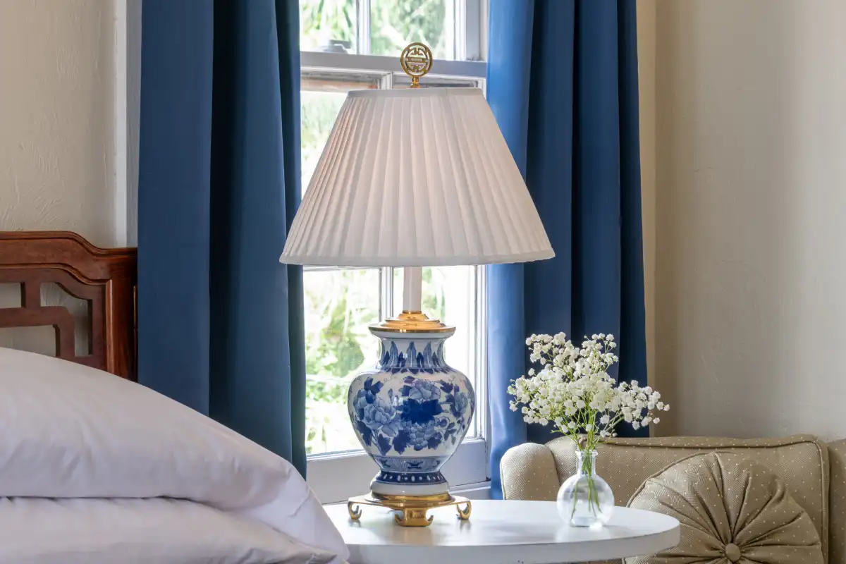 A blue and white porcelain table lamp with a pleated shade sits on a table next to blue curtains and white flowers.