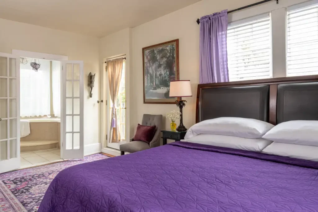 A bedroom with a purple bedspread, brown headboard, patterned rug, and open French doors leading to a bathroom.