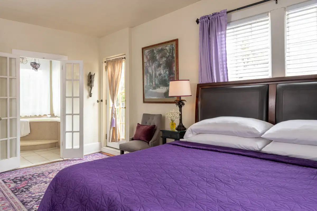 A bedroom with a purple bedspread, brown headboard, patterned rug, and open French doors leading to a bathroom.
