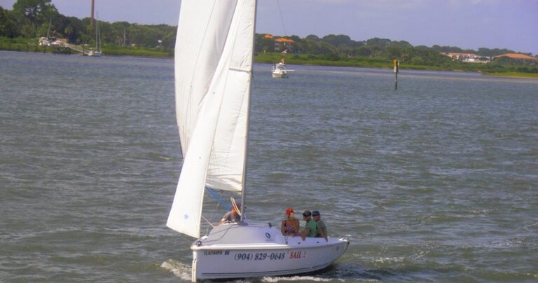 A small sailboat with a white sail carrying four people travels across a body of water toward the shore.