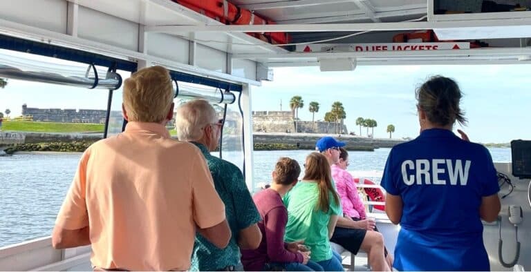 People on a boat tour look out at a shoreline with a fort in the distance, viewed from behind the crew member.