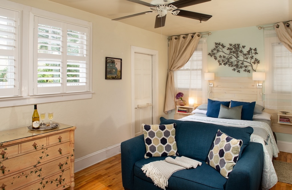 A bedroom with a bed, a blue loveseat with patterned pillows, a dresser, and two windows with white shutters.