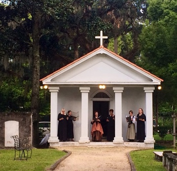 Five people in period-style clothing stand under the portico of a small white chapel with a cross on the roof.