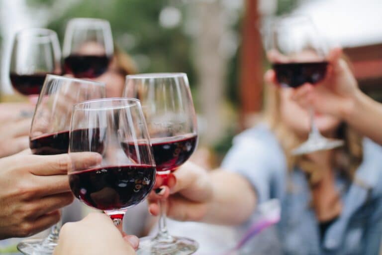 Close-up of several people holding up glasses of red wine to toast together outdoors.