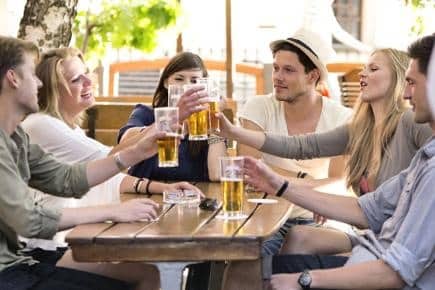 A group of young adults sit around an outdoor wooden table, smiling and clinking glasses of beer.