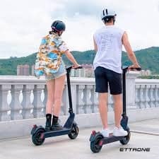 A woman and a man in helmets ride electric scooters on a walkway overlooking a distant town.