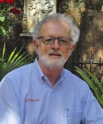 A middle-aged man with gray hair, a beard, and glasses wears a light blue button-down shirt outdoors.