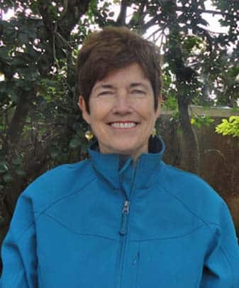A smiling woman with short brown hair wears a blue zip-up jacket against a background of trees.