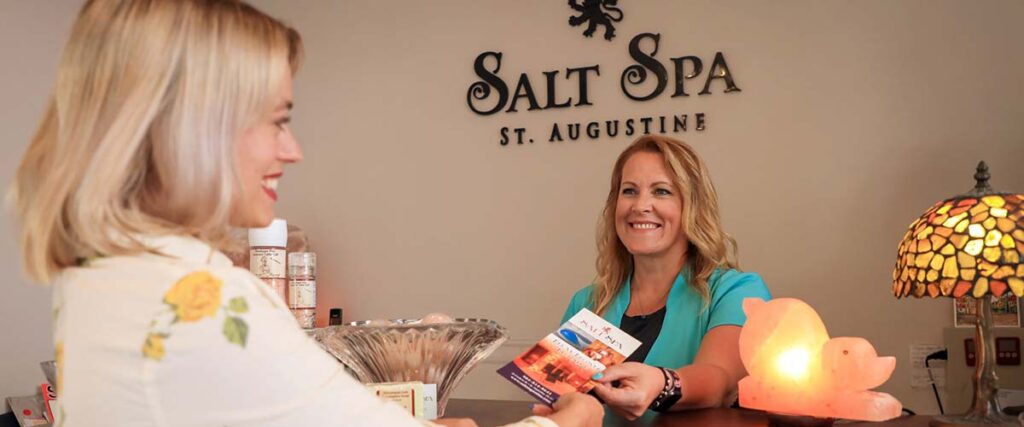 A woman hands a brochure to a receptionist at the Salt Spa St. Augustine front desk.