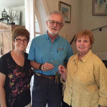 Three smiling adults standing together indoors; a man in a blue plaid shirt is flanked by two women.