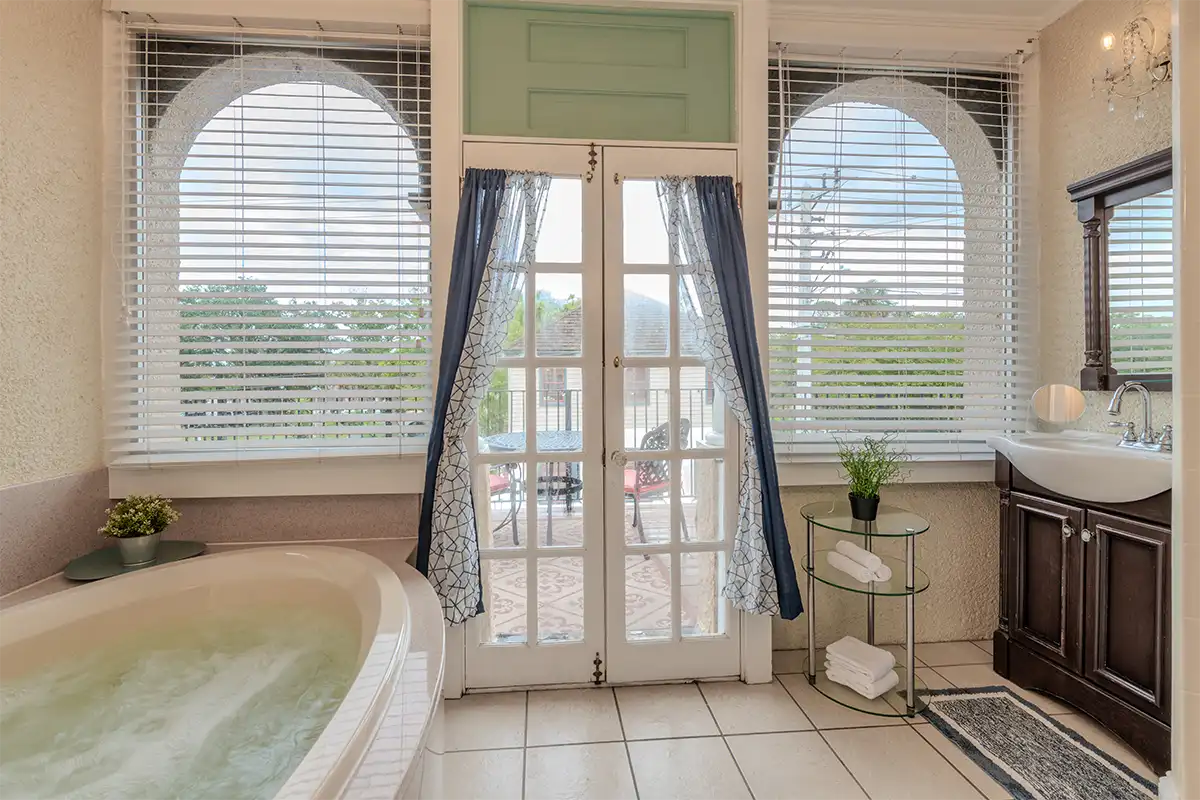 Bathroom with a soaking tub, glass doors to a balcony, and a vanity, all with white blinds on arched windows.