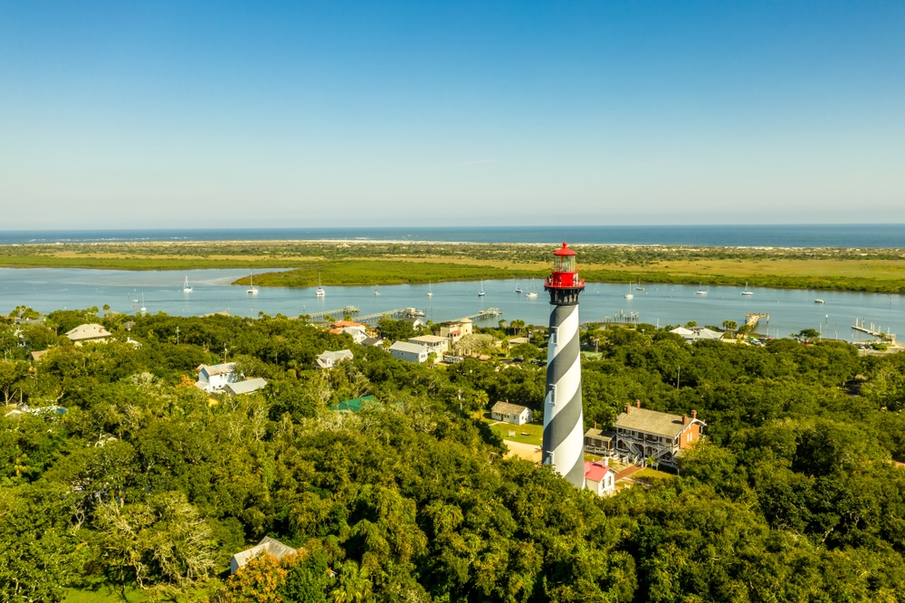 Aerial view of the St. Augustine Lighthouse surrounded by lush trees, with a bay and the ocean in the distance.