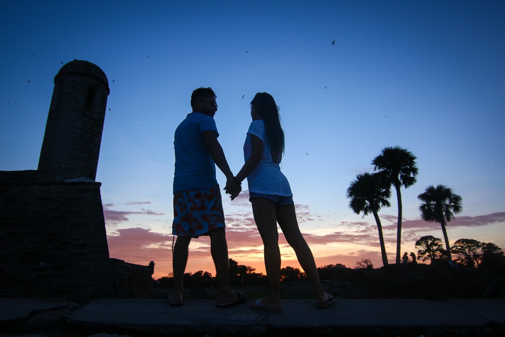 A couple holds hands in silhouette against a sunset, near a stone fort tower and palm trees.