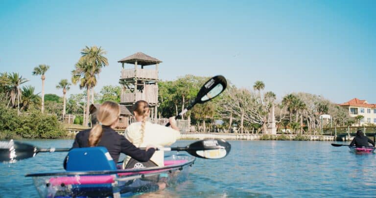 Two people in a kayak paddle on a sunny river, with a wooden structure and palm trees visible on the shore.