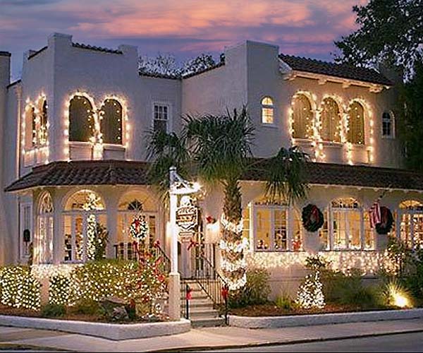 A two-story stucco building at dusk, illuminated with Christmas lights, featuring arched windows and a palm tree.