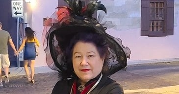 A woman wearing a large, ornate black hat with feathers poses in front of a building with a "One Way" sign.
