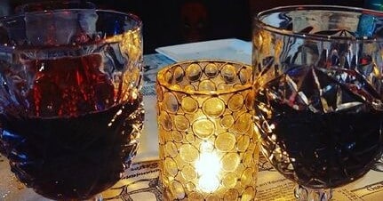 Two crystal glasses filled with dark red wine stand on either side of a glowing, textured gold candle holder.